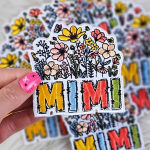May include: A white embroidered patch featuring the word "MIMI" in colourful letters surrounded by a floral design. The letters are outlined in black and have a frayed edge. The flowers are in shades of pink, yellow, and orange.
