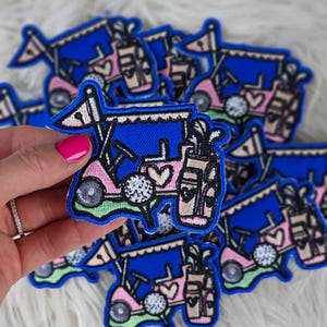 May include: Embroidered patch featuring a pink and blue golf cart with a golf ball, golf clubs, and a flag. The patch has a blue border and is decorated with hearts. The patch is held in a hand with pink nail polish.
