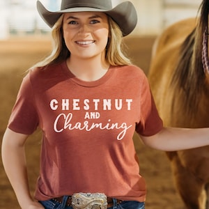 May include: A woman wearing a maroon t-shirt with the text "Chestnut and Charming" in white. She is wearing a cowboy hat and blue jeans with a silver belt buckle. She is standing next to a brown horse.