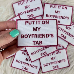 May include: White rectangular patches with claret trim and text that reads "PUT IT ON MY BOYFRIEND'S TAB" in claret thread. The patches are scattered on a white, fluffy surface. The patches are embroidered.