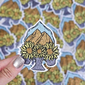 Puede incluir: Un parche bordado con una cordillera de montañas con un cielo azul, rodeado de tres girasoles amarillos. El parche tiene forma de pica y un fondo morado.