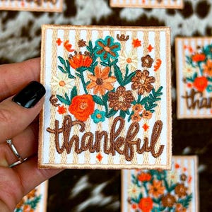 May include: A square embroidered patch featuring a white and tan vertical striped background. The design includes a colourful floral arrangement in shades of orange, teal, and brown, with the word thankful stitched in brown cursive script below.