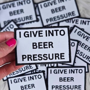Może przedstawiać: Prostokątna biała naszywka z czarną obwódką i napisem "I GIVE INTO BEER PRESSURE". Kilka podobnych naszywek jest rozrzuconych na białej, puszystej powierzchni. Tekst jest pogrubiony, bezszeryfowy.