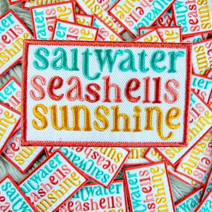 May include: Embroidered patches with the words "Saltwater, Seashells, Sunshine" in teal, coral, and yellow. The patches have a red border and are scattered on a white surface. The patches are a beach-themed product.