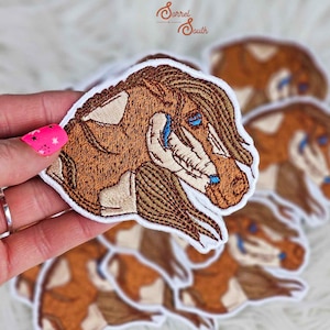 May include: A set of embroidered iron-on patches featuring a brown and white horse with blue eyes. The patches are on a white background and have a white border.