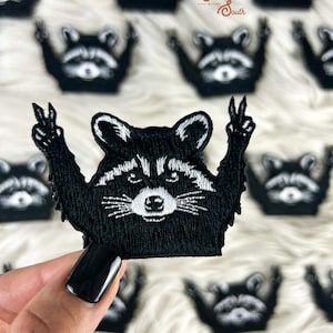 Può includere: Toppa ricamata in bianco e nero con un procione che fa il segno della pace. Il procione è nero con dettagli bianchi e alza due dita. La toppa è circondata da altre toppe con procioni. Il testo "Sorrel South" è in alto a destra.