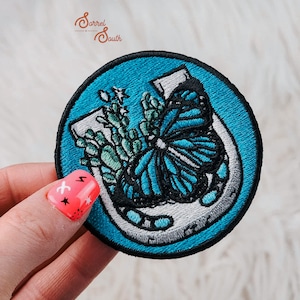 May include: A blue embroidered patch with a white horseshoe, a blue butterfly, and green foliage inside. The patch is circular and has a black outline.