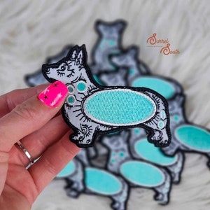 May include: A set of iron-on patches featuring a cartoon corgi with a turquoise blue oval on its back. The patches are surrounded by a black outline and have a white background.