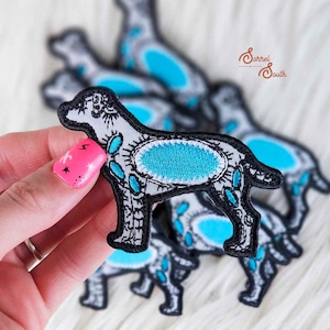 May include: Embroidered dog patch with a black outline, gray body, and turquoise accents. The patch features an oval turquoise center and decorative turquoise shapes along the legs and body. Several patches are visible.
