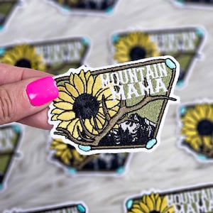 May include: Embroidered patch featuring a yellow sunflower, deer antlers, and mountain scene. The patch has the text "MOUNTAIN MAMA" in white. The patch is surrounded by a green border and light blue accents.