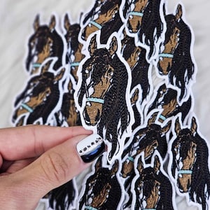 May include: A pile of brown and black horse head embroidered patches. The patches are on a white fuzzy surface.