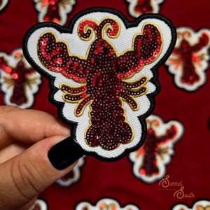 May include: A lobster-shaped patch featuring dark red sequins, gold thread embroidery, and a white fabric backing with a black border. The design includes two large claws, antennae, and multiple legs, with a repeating pattern of identical patches.