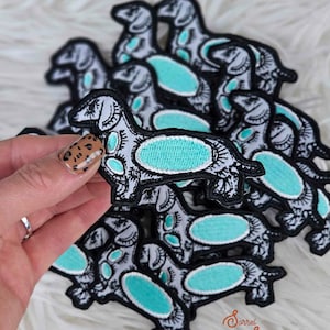 May include: A stack of embroidered iron-on patches featuring a stylized dachshund with turquoise eyes and body details. The patches are on a white fuzzy surface.