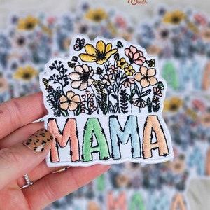 May include: A white embroidered patch with the word "MAMA" in rainbow colors. The word is surrounded by a colorful floral design.