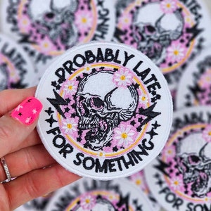 May include: White embroidered patch with a pink and yellow circle around a black and white skull with flowers. The text reads "Probably Late For Something".