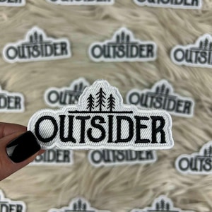 Puede incluir: Parche bordado con la palabra "OUTSIDER" en negro, contorneado en blanco, con un diseño de montaña y árboles encima del texto. Los parches están sobre una superficie suave y blanca.