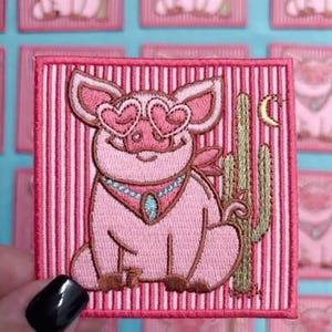 May include: A square embroidered patch featuring a pink pig wearing heart-shaped glasses and a turquoise-accented necklace. The pig sits next to a green cactus and a small yellow crescent moon on a background of vertical pink and white stripes.