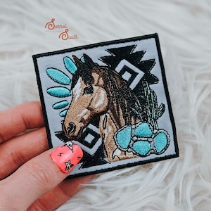 Può includere: Una toppa ricamata con la testa di un cavallo marrone e bianco con accenti turchesi. La toppa è circondata da un motivo geometrico bianco e nero. La toppa è su una superficie bianca e pelosa.