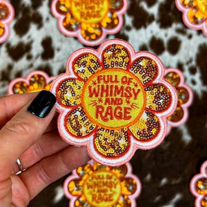 May include: A flower-shaped embroidered patch with a pink border and gold sequin petals. The center is yellow with red text that reads FULL OF WHIMSY AND RAGE. The design features a scalloped edge and a textured, stitched finish throughout the piece.