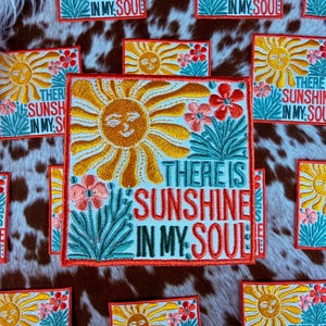 May include: Embroidered patches with a sun design and the text "THERE IS SUNSHINE IN MY SOUL". The patches feature a smiling sun, flowers, and foliage in orange, red, and teal colors. The patches are square-shaped.