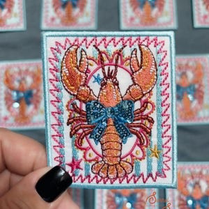 May include: A rectangular embroidered patch featuring an orange lobster centred on a white background with light blue vertical stripes. The lobster wears a blue sequined bow. The design includes small pink and yellow stars and a pink scalloped border.