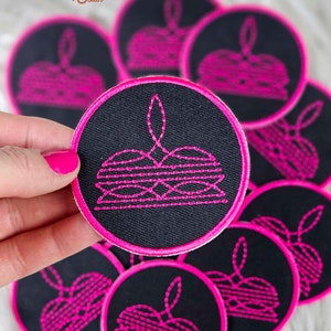 May include: A close-up of several black and pink embroidered patches. The patches are round and feature a pink design of a stack of plates. The patches are arranged in a pile.