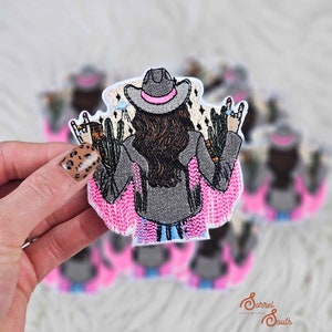 May include: Embroidered patch of a cowgirl with a grey hat and pink trim, giving a rock-and-roll hand gesture. The patch has a pink and blue fringe, cactus, and a desert landscape background.