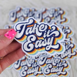 May include: A close-up of a stack of embroidered iron-on patches with a rainbow design and the text "Take it Easy" in a retro font.