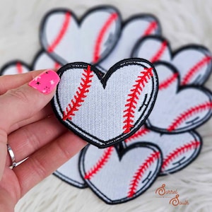 Könnte beinhalten: Herzförmige Baseball-Patches mit weißem Hintergrund und roten Nähten, schwarz umrandet. Die Patches sind so gestaltet, dass sie Basebällen ähneln. Eine Hand hält einen Patch, andere im Hintergrund.
