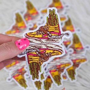 May include: A set of embroidered iron-on patches featuring a pair of yellow, pink, and brown sneakers with a green cactus on top. The patches are on a white fuzzy surface.
