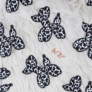 May include: A collection of black and white dalmatian-patterned balloon dog patches on a white, fluffy background. The patches, shaped like balloon animals, have a black border and a pattern of black spots on a white base. These patches are designed for application to fabric.