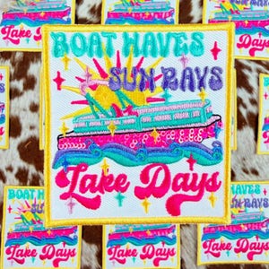 以下が含まれることがあります： 鮮やかな色で「Boat Haves + Sun Rays」と「Lake Days」のフレーズが刺繍されたパッチ。中央のパッチは、太陽光線のあるボートのデザインが特徴です。パッチは金色の縁取りのある白い背景にあります。