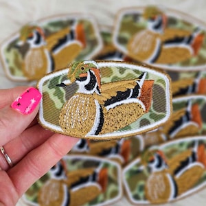 May include: Embroidered patch featuring a detailed wood duck design. The duck has a colorful head, brown body, and orange accents. The patch is bordered by a white stitch and set against a camouflage background.
