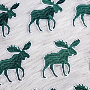 Puede incluir: Parches bordados con un diseño de alce verde con contornos blancos. Los parches están dispuestos sobre una superficie blanca y esponjosa. El alce tiene cuernos detallados y una pose de caminar, para ropa o accesorios.