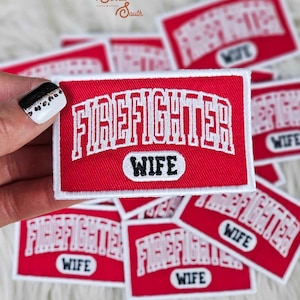 Pode incluir: Patches bordados vermelhos e brancos com a inscrição "FIREFIGHTER WIFE". O texto branco destaca-se no fundo vermelho. Vários patches são visíveis, com uma borda branca.