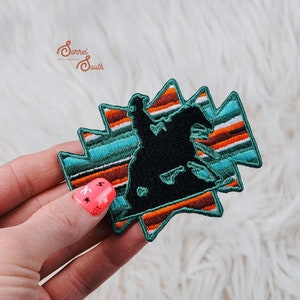 May include: A black silhouette of a person riding a horse, embroidered on a turquoise, orange and white striped fabric patch. The patch is shaped like a starburst.