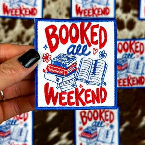 May include: A rectangular embroidered patch with a white background and a blue border. The text reads BOOKED all WEEKEND in red and blue. The design features a stack of three books and an open book with blue and red thread details.