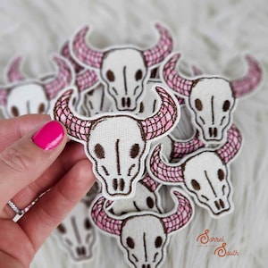 May include: Embroidered patches featuring a white skull with brown details and pink plaid horns. The patches are in a pile, with one held by a hand with pink nail polish. The patches are likely for clothing or accessories.