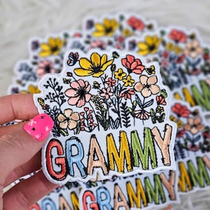 Puede incluir: Un parche blanco bordado con un diseño floral colorido y la palabra "GRAMMY" en letras de arcoíris.