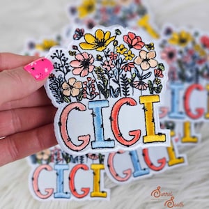 May include: A set of embroidered iron-on patches with the word "GIGI" in colorful letters surrounded by a bouquet of flowers. The patches are on a white background.