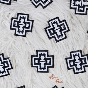 Può includere: Toppe ricamate in bianco e nero con un design a croce geometrica. Le toppe sono disposte su una superficie bianca e soffice. Il design presenta un motivo centrale con motivi simmetrici e un bordo nero.