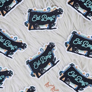 May include: A collection of nine black and white embroidered patches featuring a cow silhouette with the text "Eat Beef" in turquoise blue. The patches are on a white fluffy surface.