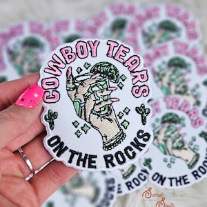 May include: A white embroidered patch with pink lettering that reads "Cowboy Tears on the Rocks". The patch features a hand holding a margarita glass with a lime wedge. There are green cacti and stars around the glass.
