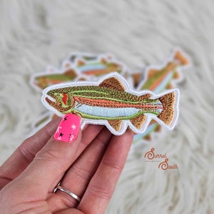 May include: A colorful embroidered iron-on patch of a trout fish. The patch is surrounded by a white border and has a brown, green, orange, and blue color scheme. The patch is held in a hand with pink nail polish.