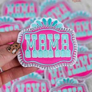 Puede incluir: Primer plano de un parche bordado de color rosa y turquesa con la palabra "MAMA" en una fuente de estilo vintage. El parche tiene un borde blanco y un diseño turquesa decorativo en la parte superior.