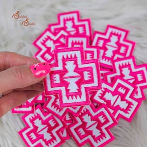 Op de afbeelding: Een stapel roze en witte geborduurde patches met een geometrisch ontwerp. De patches zijn in de vorm van een kruis.