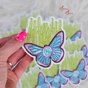 May include: Embroidered patch with a butterfly design, featuring teal and purple wings and a brown outline. The patch has a green background and a white border. The patch is held in a hand. Suitable for clothing or accessories.