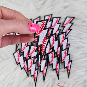 May include: Baseball-themed lightning bolt patches. Each patch features a white background with red stitching, mimicking a baseball. The edges are outlined in black. The patches are arranged on a fluffy, white surface.