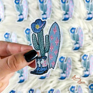 Può includere: Toppa ricamata raffigurante un cactus con cappello e stivali da cowboy, con una tavola da surf. Il cactus è verde, il cappello e gli stivali sono blu e la tavola da surf è azzurra con cactus rosa. Sfondo bianco e soffice.