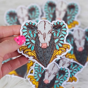 May include: A close-up of an embroidered iron-on patch featuring a cow's head with a white face and black and gray horns. The cow's head is surrounded by turquoise blue and gold leaves. The patch is on a white background.
